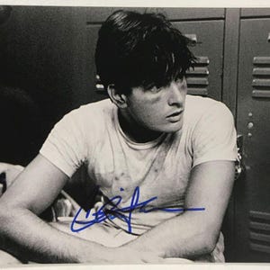 May include: Black and white photograph of a man in a white t-shirt, seated with arms crossed. The image is signed in blue ink. The background features a series of gray lockers.