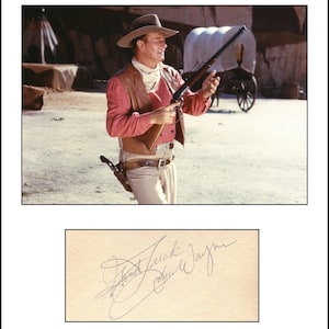 Peut inclure: Une photographie de John Wayne portant un chapeau de cow-boy beige, une chemise rouge à manches longues, un gilet en cuir marron et un étui avec un revolver, tenant un fusil. Sous l'image, une carte blanche porte la signature Good Luck John Wayne.
