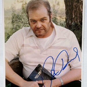 May include: A signed photograph featuring a man in a light-colored collared shirt, holding a book labeled "Holy Bible." The photo has a blue autograph and a background of greenery.