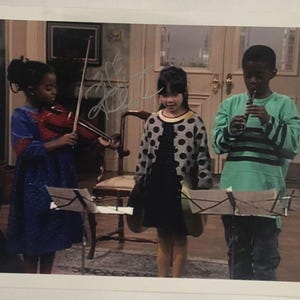 May include: A photograph of three children playing musical instruments. A girl in a blue dress plays a violin, another girl wears a black dress and a polka-dot sweater, and a boy plays a flute. Music stands are in front of them.