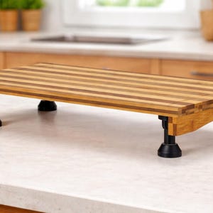 May include: A rectangular bamboo cutting board with a striped pattern, supported by four black legs. The board is elevated, designed for kitchen use. The wood tones are light and dark brown.