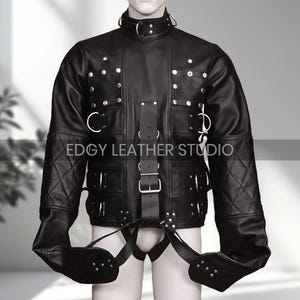 Puede incluir: Chaqueta de cuero negro con cuello alto, múltiples correas y herrajes metálicos. La chaqueta presenta detalles de costuras de diamante en las mangas y está diseñada con restricciones de brazo adjuntas. El texto "EDGY LEATHER STUDIO" es visible.