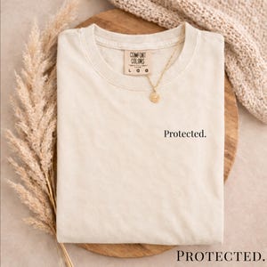 Może przedstawiać: Kremowa koszulka Comfort Colors z napisem "Protected" z przodu. Złoty naszyjnik z wisiorkiem spoczywa na koszulce. Koszulka jest złożona na drewnianej powierzchni.