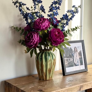 May include: A decorative display featuring a vase of magenta peonies and blue delphiniums. The vase is green and brown. A framed photograph sits on a wooden table, creating a homely feel.