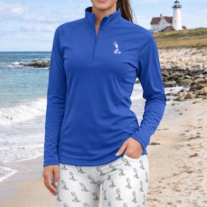 May include: A woman wearing a royal blue long-sleeved top with a small white graphic and white leggings patterned with champagne glasses. A lighthouse is visible in the background. The outfit is suitable for outdoor activities.