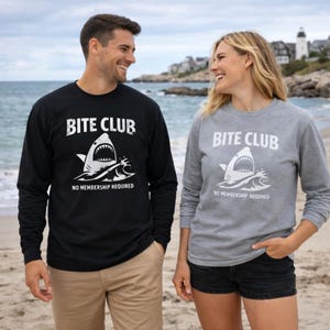 May include: Black and gray long-sleeve shirts with the text "BITE CLUB" above a shark graphic. The shark graphic has an open mouth with sharp teeth. Below the graphic is the text "NO MEMBERSHIP REQUIRED".
