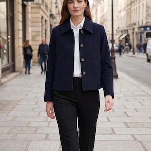 May include: A woman wearing a navy blue jacket over a white button-down shirt and black pants. The jacket has a collar and button closure. The outfit is classic and stylish, suitable for a professional or casual setting. The background shows a city street.