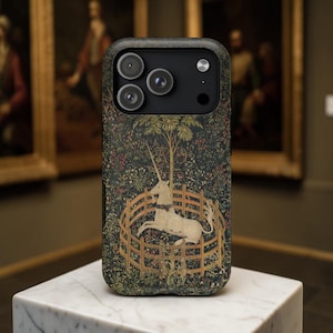 May include: A phone case featuring a tapestry design with a white unicorn inside a wooden fence, surrounded by a forest. The case has a dark background with a textured appearance and a black camera module.