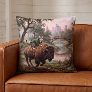 May include: A square throw pillow featuring a printed illustration of a tabby cat wearing a green jacket while riding a brown bison. The scene includes a river, a wooden arched bridge, and a distant city skyline under a soft, hazy sky.