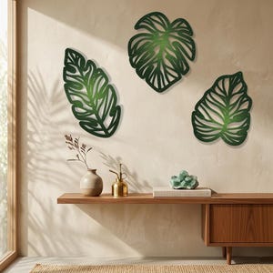 May include: Three green metal leaf wall art pieces in varying shapes are mounted on a neutral wall. The leaves are cut out with detailed vein patterns. A wooden shelf below holds a vase, a mister, and a decorative crystal.