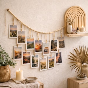 Peut inclure: Présentoir mural avec photos de paysages, suspendues à une guirlande de perles en bois et de petites pinces en bois. Les photos montrent des couchers de soleil et des paysages désertiques. Une étagère en bois avec des objets décoratifs est également visible.