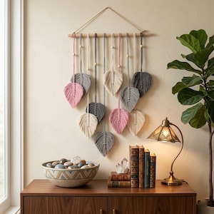 May include: A macrame wall hanging with leaf-shaped designs in pink, grey, and cream. The leaves are suspended from a wooden dowel with cords and wooden beads. This wall decor piece adds a touch of bohemian style to any room.