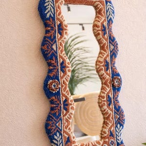 May include: A decorative wall mirror with a distinctive, textured frame. The frame features a wavy design in blue, brown, and cream, with detailed patterns. The mirror's design evokes a textile or rug, offering a unique decorative touch for any room.