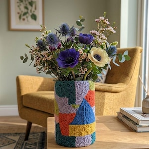 May include: A colorful, patterned vase holds a vibrant bouquet of flowers. The vase features a geometric design with patches of blue, orange, yellow, and purple. The flowers include purple anemones, blue thistles, and delicate pink and white blossoms.