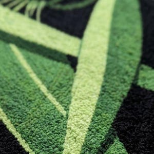 May include: Close-up of a textured, green and black rug. The design features large, stylized green leaves against a black background. The varying shades of green and the texture create a sense of depth and visual interest. The rug appears to be made of a soft, looped material.