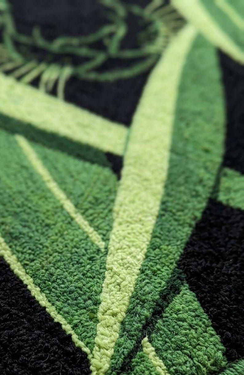 May include: Close-up of a textured, green and black rug. The design features large, stylized green leaves against a black background. The varying shades of green and the texture create a sense of depth and visual interest. The rug appears to be made of a soft, looped material.