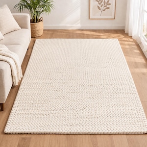 Könnte beinhalten: Ein großer, rechteckiger, cremefarbener Teppich mit einem dicken, strukturierten Gewebe. Der Teppich liegt auf einem Holzboden in einem Wohnzimmer, mit einem Sofa, einer Decke und einer Topfpflanze im Hintergrund. Die neutrale Farbe des Teppichs ergänzt die Einrichtung des Raumes.