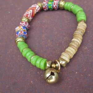 以下が含まれることがあります： 緑、茶色、赤のビーズで作られたブレスレットで、金色のベルのチャームが付いています。ブレスレットは、木、ガラス、金属など、さまざまな種類のビーズで作られています。