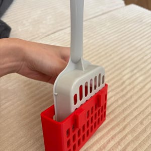 Può includere: Una paletta per lettiera per gatti in plastica grigia e bianca con un contenitore rosso. La paletta ha un design a fessura e un lungo manico. Il contenitore rosso ha una forma rettangolare con più fori.
