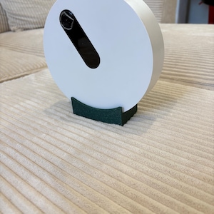 Peut inclure: Un objet circulaire blanc avec une ouverture oblongue noire, monté sur une base vert foncé. L'objet repose sur une surface beige texturée en velours côtelé. Le design est moderne et minimaliste.