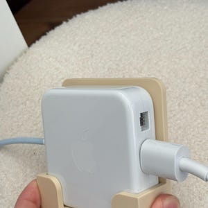 Puede incluir: Un adaptador de corriente Apple blanco con el logotipo de Apple, sujeto en un soporte beige. El adaptador tiene un cable de carga blanco enchufado. El soporte es de color beige claro.