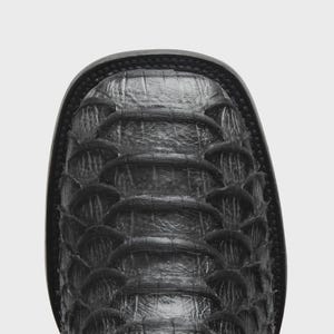 May include: A black square-toe boot featuring a textured, overlapping scale-like pattern across the vamp. The boot has a dark, matte finish with visible horizontal ridges and a thick, stitched welt surrounding the perimeter of the toe box.