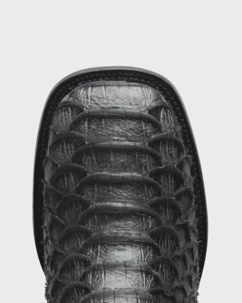 May include: A black square-toe boot featuring a textured, overlapping scale-like pattern across the vamp. The boot has a dark, matte finish with visible horizontal ridges and a thick, stitched welt surrounding the perimeter of the toe box.