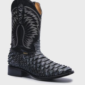 May include: A black cowboy boot featuring a textured, scale-like pattern on the vamp and heel counter. The tall shaft is made of smooth black leather with intricate white decorative stitching in a wing-like design and a square toe profile.