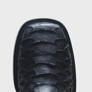 May include: A black boot with a square toe and a textured, overlapping scale-like pattern across the vamp. The boot features a thick, dark sole with visible stitching along the edge, showcasing a distinct, layered material construction.