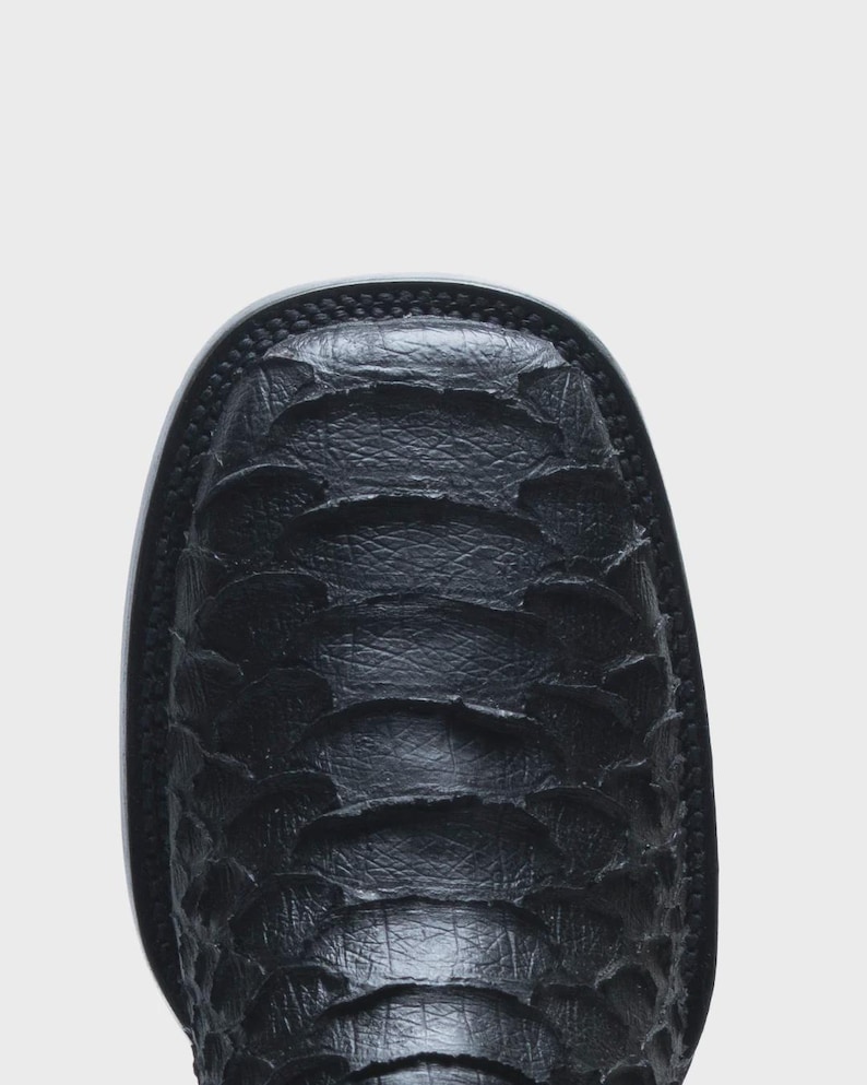 May include: A black boot with a square toe and a textured, overlapping scale-like pattern across the vamp. The boot features a thick, dark sole with visible stitching along the edge, showcasing a distinct, layered material construction.