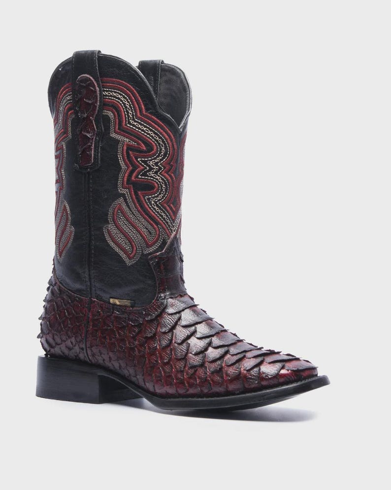 May include: A mid-calf western boot featuring a dark red, textured scale-patterned vamp and a black leather shaft. The shaft is decorated with intricate red and white stitched embroidery patterns and includes black pull tabs at the top opening.