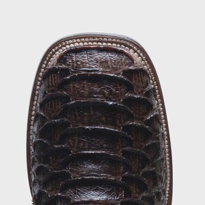 May include: A dark brown square-toe boot featuring a textured, overlapping scale pattern across the vamp. The boot is constructed with visible light-colored stitching along the welt, providing a clear contrast against the dark leather finish.