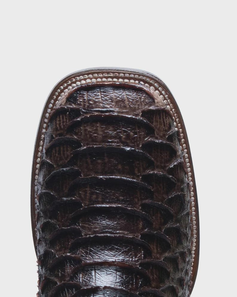 May include: A dark brown square-toe boot featuring a textured, overlapping scale pattern across the vamp. The boot is constructed with visible light-colored stitching along the welt, providing a clear contrast against the dark leather finish.