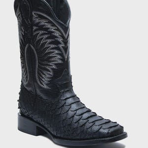 May include: A black western-style boot featuring a textured, scale-like pattern on the foot and a smooth leather shaft. The upper shaft is decorated with intricate white stitching in a wing-like design, complete with pull tabs at the top.