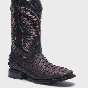 Pode incluir: Uma bota western preta com um padr&atilde;o texturizado semelhante a escamas no p&eacute; com tons avermelhados. O cano alto exibe costuras decorativas intrincadas em branco e vermelho com design de asas, com puxadores e um salto bloco.
