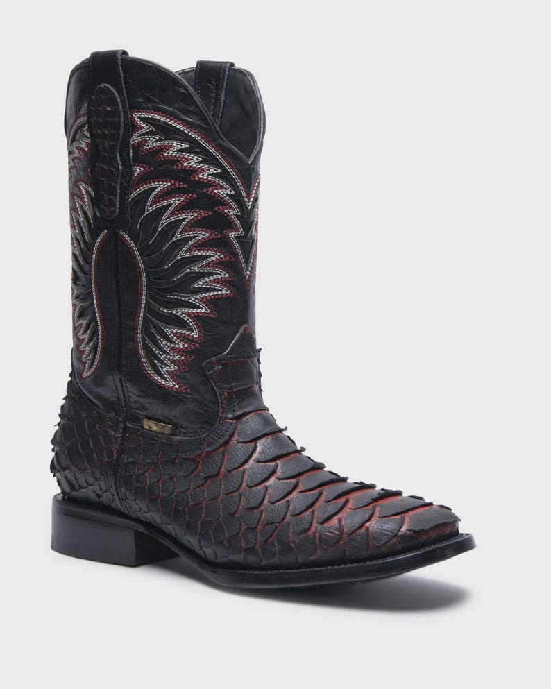 Pode incluir: Uma bota western preta com um padr&atilde;o texturizado semelhante a escamas no p&eacute; com tons avermelhados. O cano alto exibe costuras decorativas intrincadas em branco e vermelho com design de asas, com puxadores e um salto bloco.