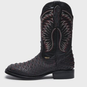 Pode incluir: Uma bota de cowboy preta com um padr&atilde;o texturizado de escamas no peito do p&eacute; e no calcanhar, com detalhes vermelhos subtis entre as escamas. O cano alto apresenta costuras decorativas complexas em branco e vermelho e duas abas no topo.