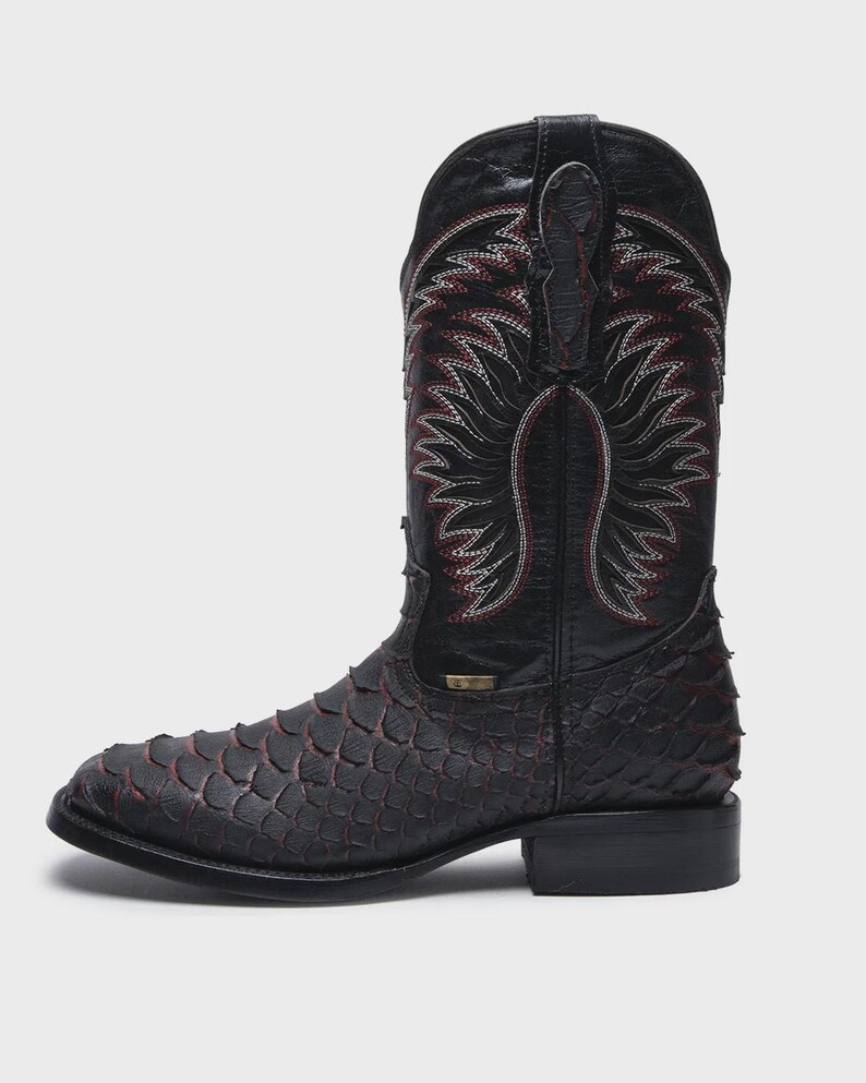 Pode incluir: Uma bota de cowboy preta com um padr&atilde;o texturizado de escamas no peito do p&eacute; e no calcanhar, com detalhes vermelhos subtis entre as escamas. O cano alto apresenta costuras decorativas complexas em branco e vermelho e duas abas no topo.
