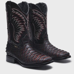 Pode incluir: Um par de botas de cowboy em couro preto com um padr&atilde;o texturizado de escamas no p&eacute; e detalhes em vermelho. Os canos altos exibem costuras decorativas intrincadas em branco e vermelho, bico quadrado e salto bloco para um perfil cl&aacute;ssico.