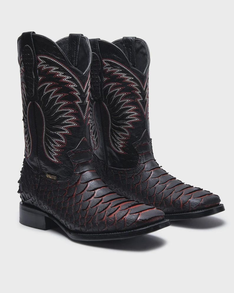 Pode incluir: Um par de botas de cowboy em couro preto com um padr&atilde;o texturizado de escamas no p&eacute; e detalhes em vermelho. Os canos altos exibem costuras decorativas intrincadas em branco e vermelho, bico quadrado e salto bloco para um perfil cl&aacute;ssico.