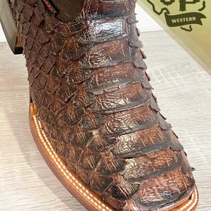 May include: A brown western boot featuring a textured, scale-like pattern across the vamp and toe. The boot has a square toe, a stacked heel, and white contrast stitching along the welt. A logo reading JP WESTERN is visible in the corner.