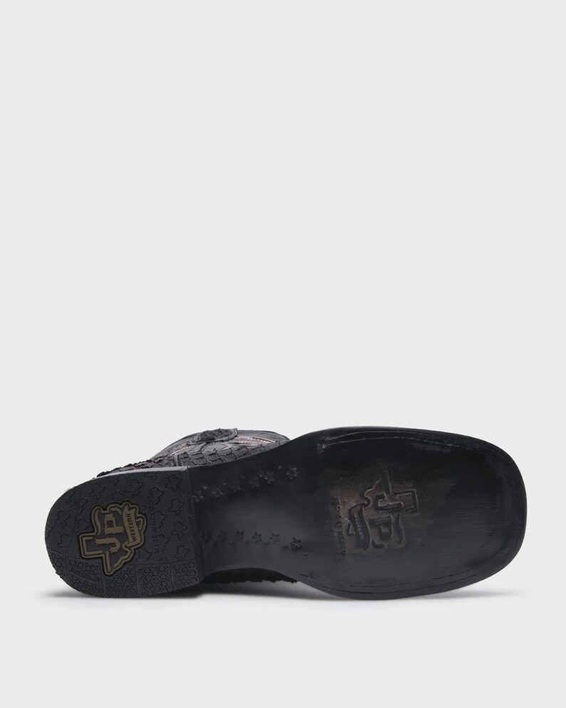 May include: A black leather western boot with a square toe and a stacked heel. The sole features a stamped logo with the letters JP inside a state of Texas outline, surrounded by a repeating star pattern and additional embossed branding.