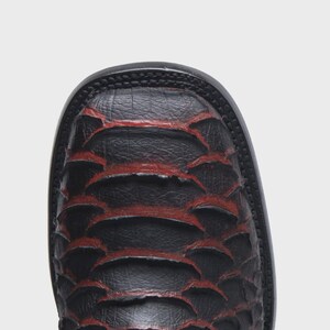 Pode incluir: Uma bota de bico quadrado com acabamento preto texturizado e detalhes em vermelho profundo vis&iacute;veis entre os padr&otilde;es sobrepostos em forma de escamas. A bota possui uma sola preta grossa e um perfil estruturado de bico quadrado arredondado.