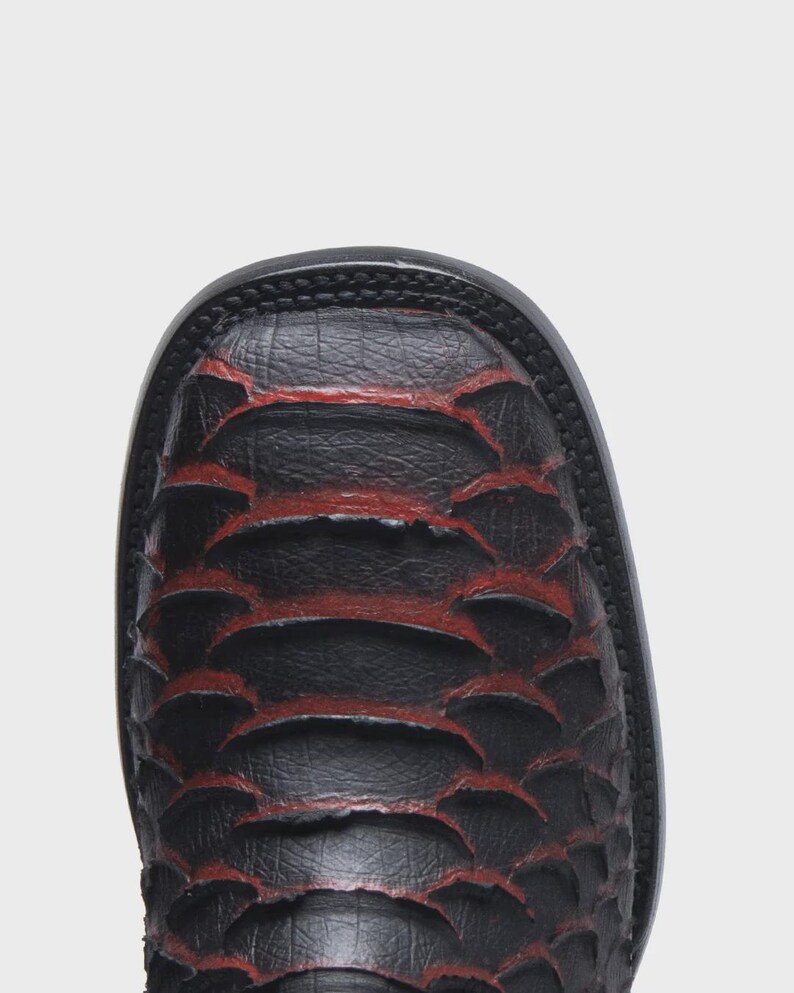 Pode incluir: Uma bota de bico quadrado com acabamento preto texturizado e detalhes em vermelho profundo vis&iacute;veis entre os padr&otilde;es sobrepostos em forma de escamas. A bota possui uma sola preta grossa e um perfil estruturado de bico quadrado arredondado.