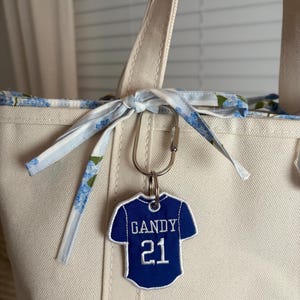 May include: A cream-colored canvas tote bag with a blue and white floral fabric tie at the top. A blue jersey-shaped keychain with white embroidery reading GANDY and the number 21 is attached to the bag with a silver metal carabiner.