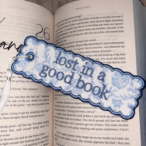 May include: A rectangular fabric bookmark with scalloped edges, featuring a light blue and white floral pattern. The text "lost in a good book" is embroidered in dark blue thread across the center. A white tassel is attached to the left side.