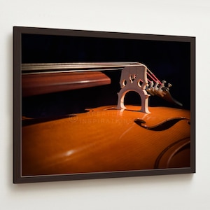 May include: Close-up of a framed photograph of a cello. The image highlights the instrument's wooden body, bridge, strings, and scroll. The cello's warm, brown tones contrast with the dark background, creating a dramatic effect. The image is framed in a dark brown frame.