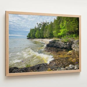 May include: A framed landscape photograph of a shoreline with a rocky coast, green trees, and a cloudy sky. The water has small waves. The frame is made of light-coloured wood.