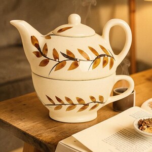 Puede incluir: Juego de tetera y taza de cerámica color crema con diseño de hojas marrones. La tetera se asienta sobre la taza, creando una apariencia apilada. El vapor se eleva de la tetera, lo que sugiere té caliente. El juego está sobre una mesa de madera.