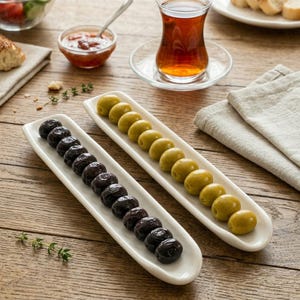 Peut inclure: Une table en bois avec deux plats de service rectangulaires blancs remplis d'olives. Un plat contient des olives noires, l'autre des olives vertes. Un verre de thé et de petits bols de nourriture sont également présents.